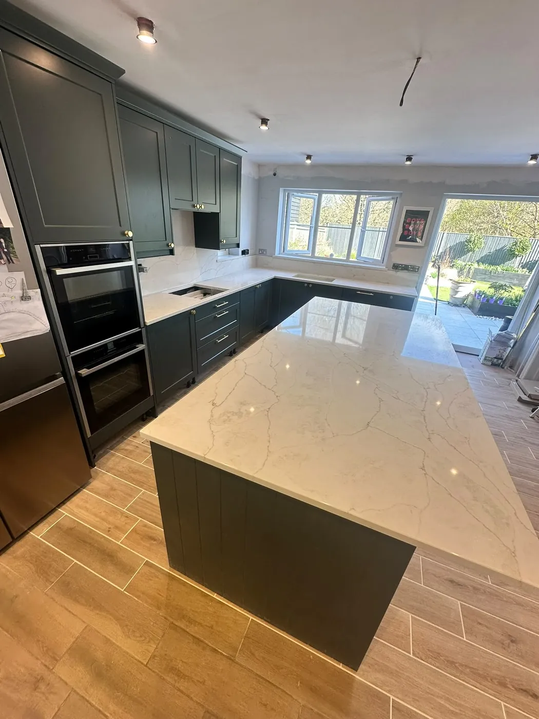 Dark green shaker kitchen we fitted, with a large marble-effect island, double oven tower and wood-effect tiled floor