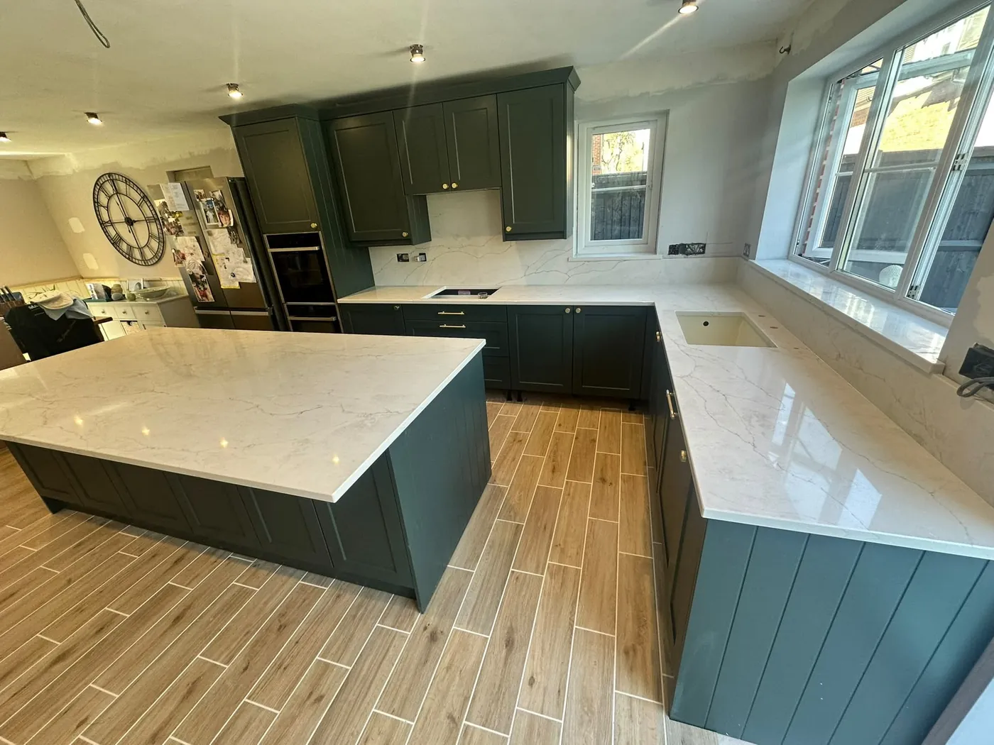 Dark green shaker kitchen with a marble-effect island and worktops running under the window